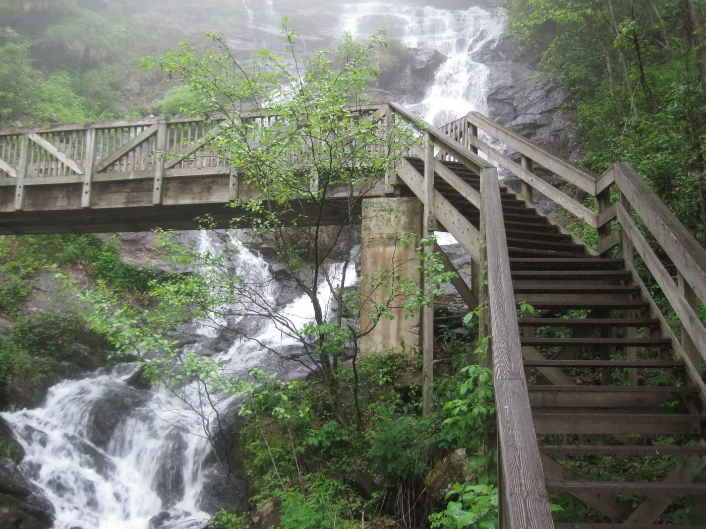 A wooden footbridge with braced railings spanning a misty cascade, with stairs climbing to the right