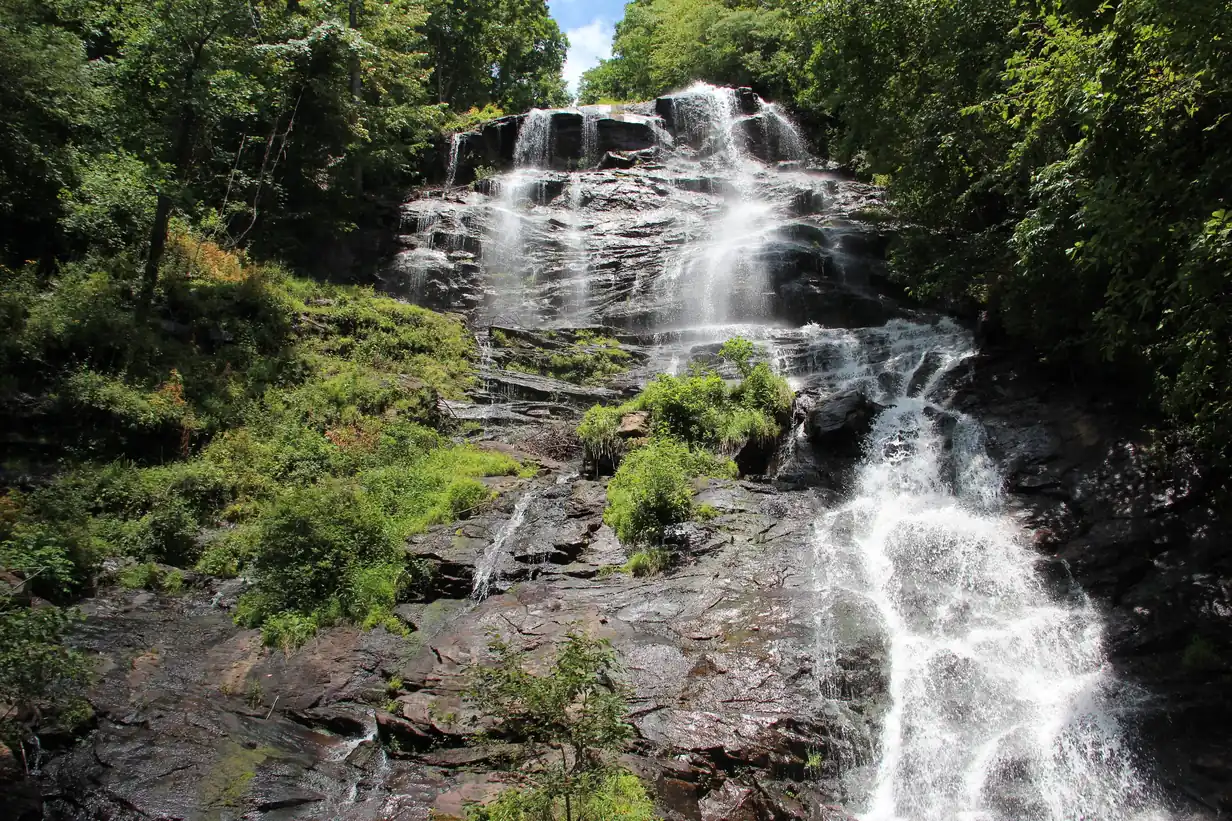 A tall waterfall fanning down a dark stepped rock face between dense green forest