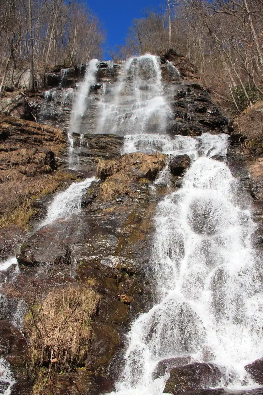 The full height of a waterfall spreading in white sheets over brown and black rock beneath bare grey trees