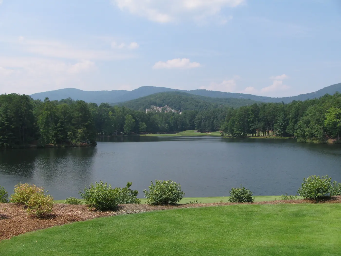 A dark still lake with a golf fairway on the far shore and layered blue ridges behind