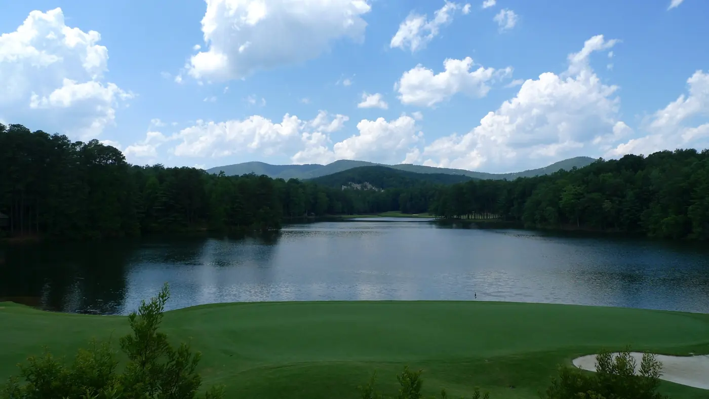 A mountain lake under a bright sky of white clouds, with a golf green along the near shore
