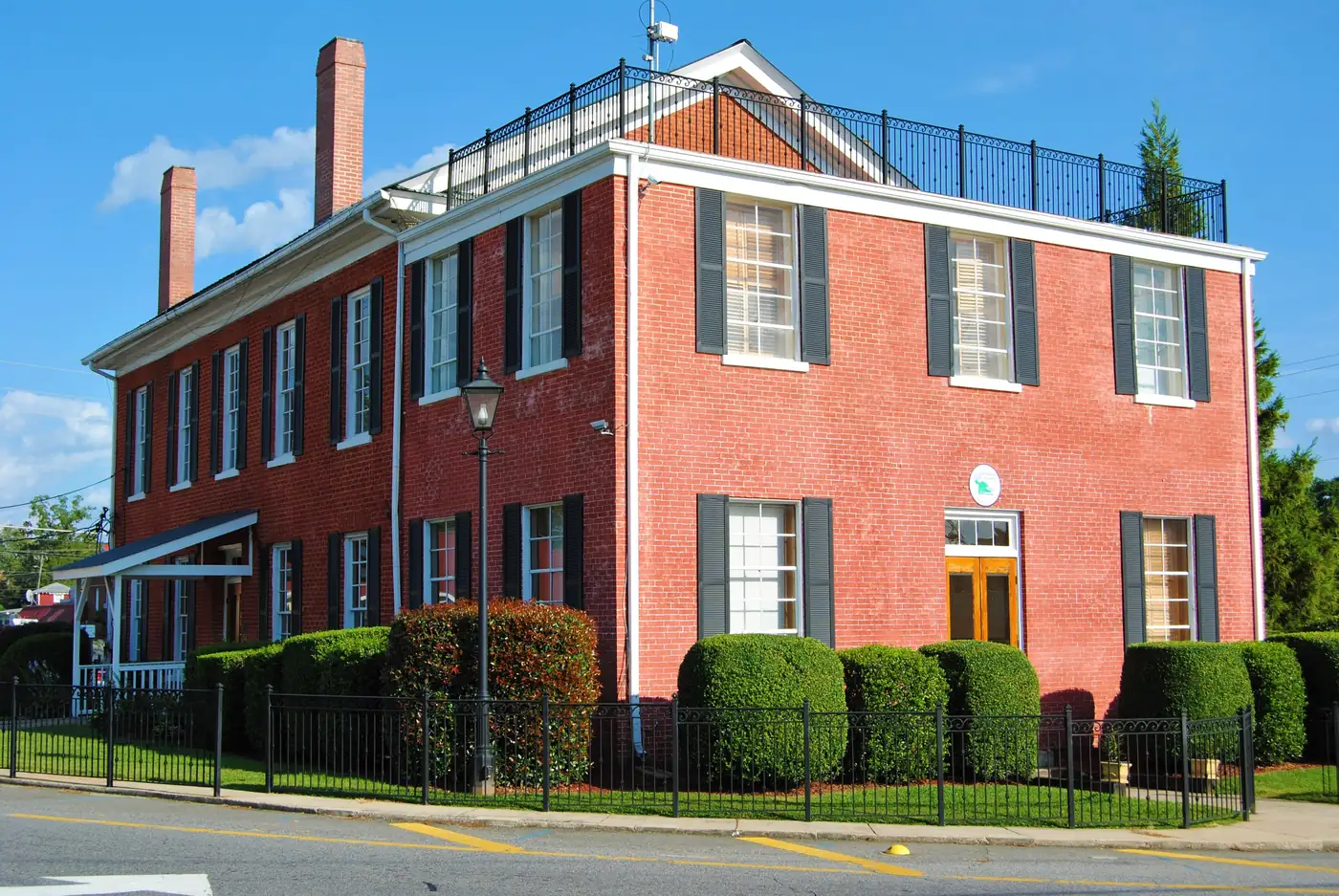A two-storey red-brick building with black shutters, two chimneys and a white cornice behind an iron fence and clipped shrubs