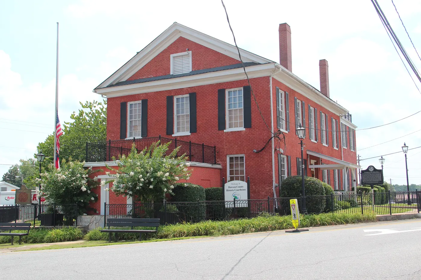 A red-brick historic courthouse with a flagpole, white crape myrtles in bloom and a welcome sign in front