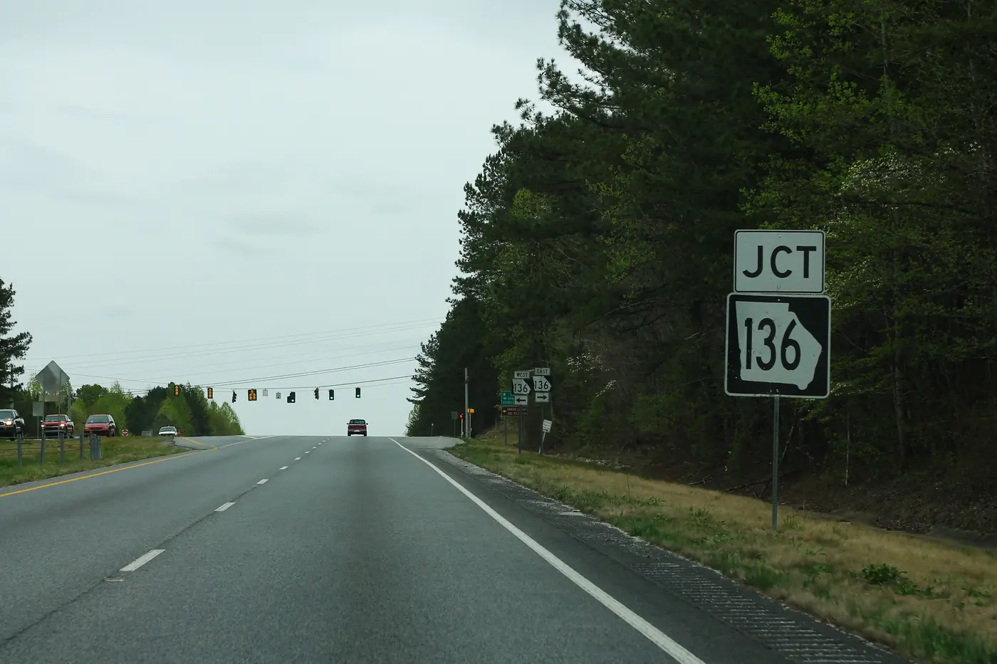 A four-lane divided highway with a large JCT 136 route sign on the shoulder in front of pine woods