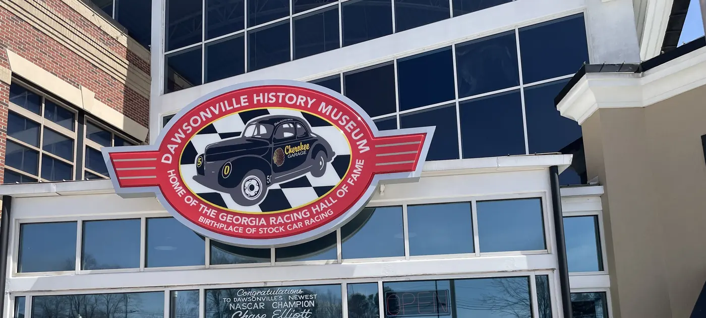 An oval sign with a vintage stock car reading Dawsonville History Museum, Home of the Georgia Racing Hall of Fame, on a glass wall