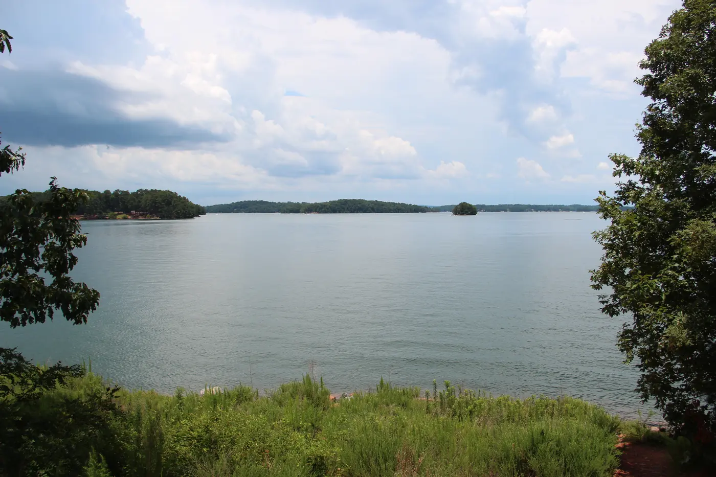 A wide calm lake with low wooded islands under towering white summer clouds, framed by leafy trees