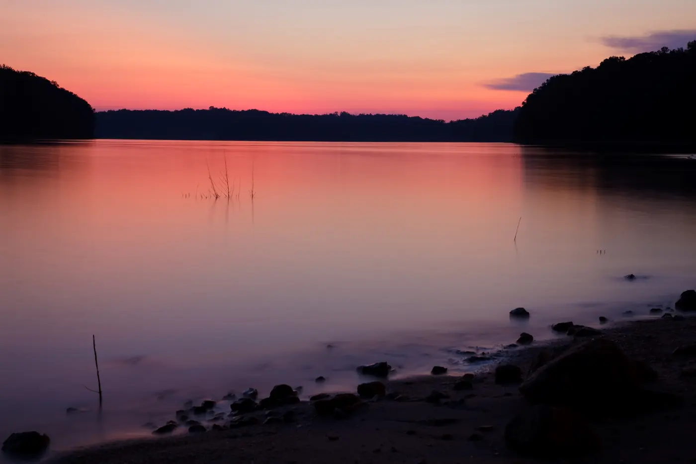 A pink and orange sunset reflected in a glassy lake beneath a dark wooded shoreline