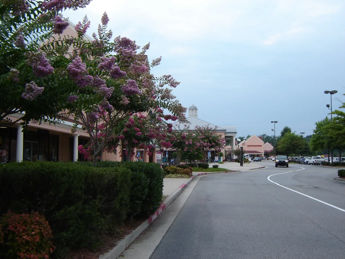 A curving drive past pink outlet storefronts with white columns, lined with crape myrtles in bloom