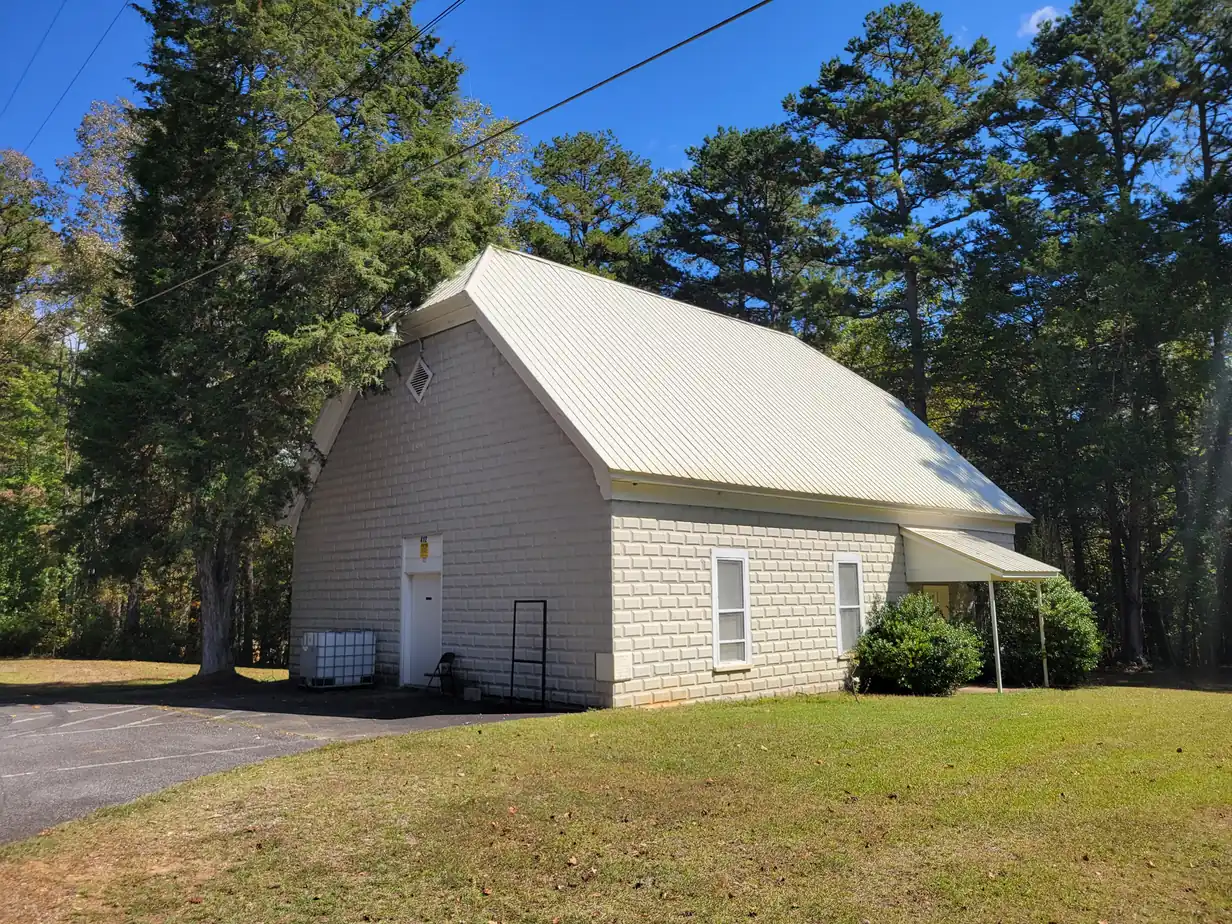 A small white country church with a cream metal gable roof among tall pines and cedars
